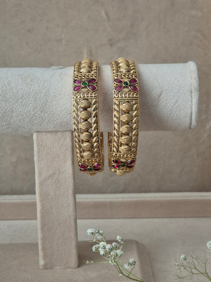 Pair of Naitri's Zahira gold bangles showcasing detailed antique-style craftsmanship, embedded with vibrant pinkish-red floral motifs and green accent stones, by naitri.in