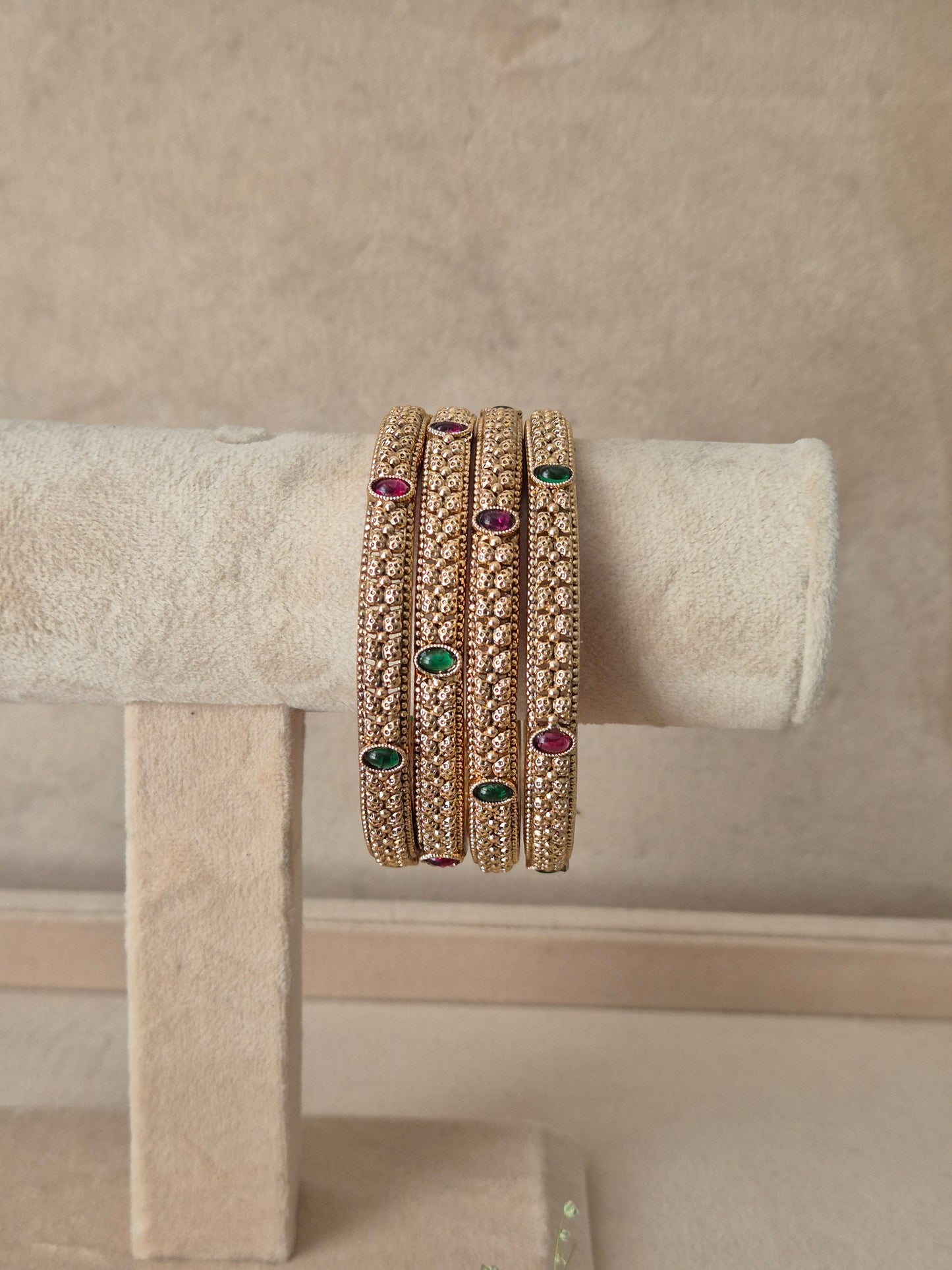Set of four gold-toned bangles featuring detailed texture with alternating ruby-red and emerald-green oval gemstone accents, by naitri.in