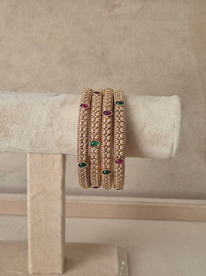Set of four gold-toned bangles featuring detailed texture with alternating ruby-red and emerald-green oval gemstone accents, by naitri.in