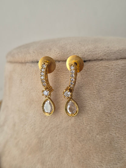 A pair of gold drop earrings with small clear stones along the curved top bar and a larger clear teardrop-shaped stone at the bottom, by naitri.in