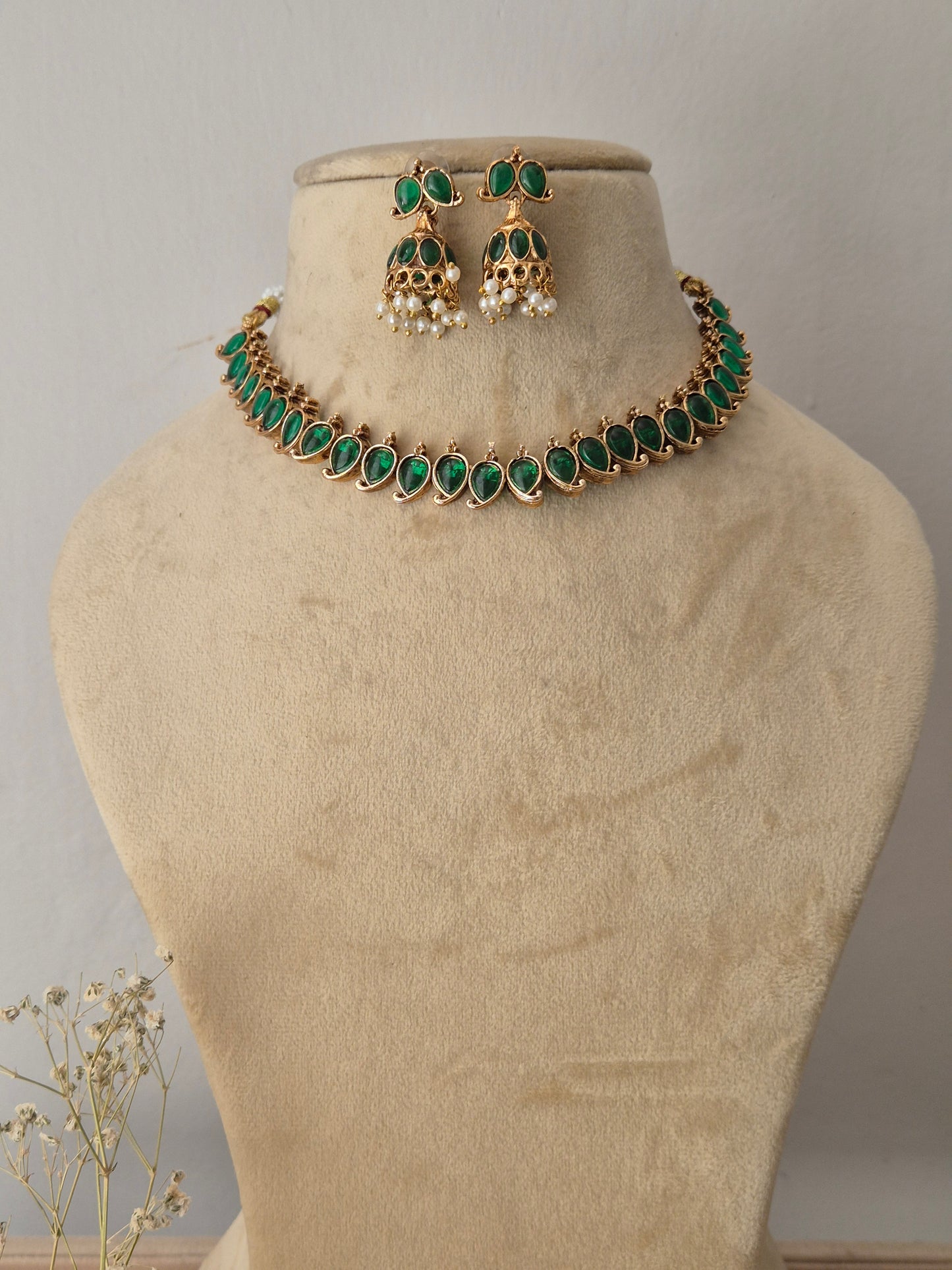 Gold-toned traditional choker necklace and matching dangle earrings, choker features a row of vibrant oval-cut green gemstones bordered by tiny gold accents, paired with matching bell-shaped jhumka-style earrings embellished with green stones and hanging white pearl droplets by naitri.in