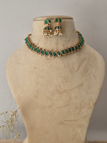 Gold-toned traditional choker necklace and matching dangle earrings, choker features a row of vibrant oval-cut green gemstones bordered by tiny gold accents, paired with matching bell-shaped jhumka-style earrings embellished with green stones and hanging white pearl droplets by naitri.in
