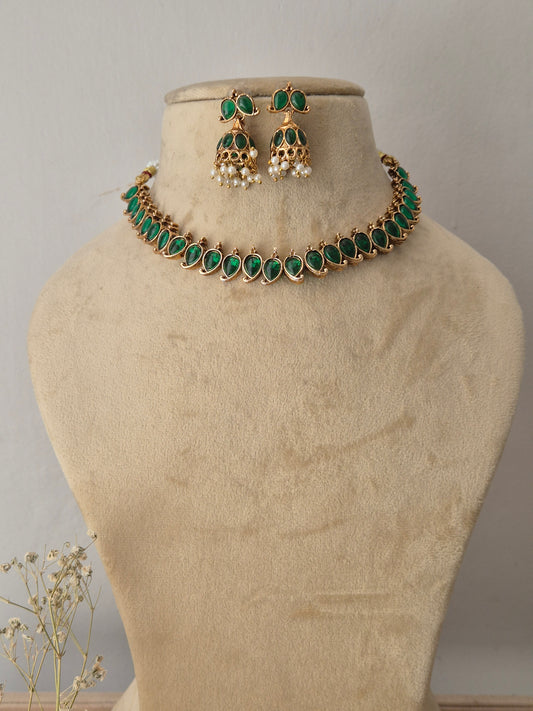 Gold-toned traditional choker necklace and matching dangle earrings, choker features a row of vibrant oval-cut green gemstones bordered by tiny gold accents, paired with matching bell-shaped jhumka-style earrings embellished with green stones and hanging white pearl droplets by naitri.in