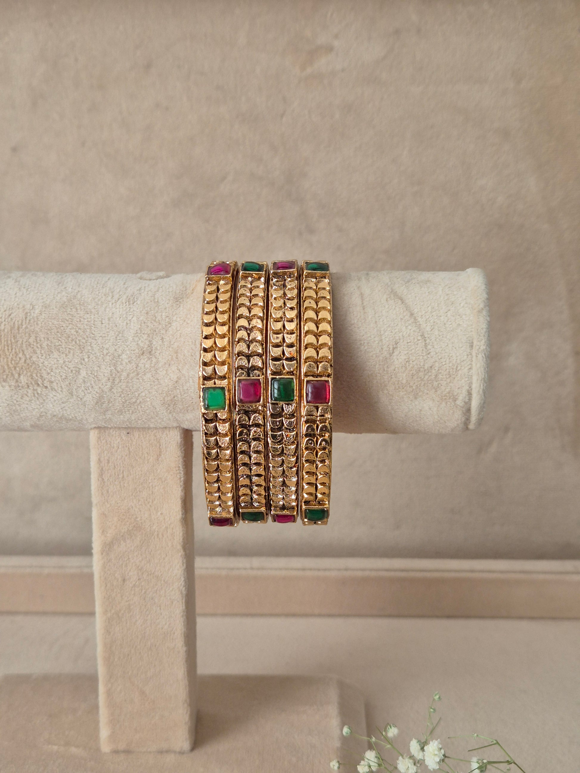 Four stacked gold bangles with textured patterns and rectangular green and pink stone accents, by naitri.in
