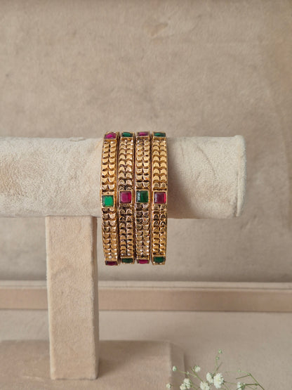 Four stacked gold bangles with textured patterns and rectangular green and pink stone accents, by naitri.in