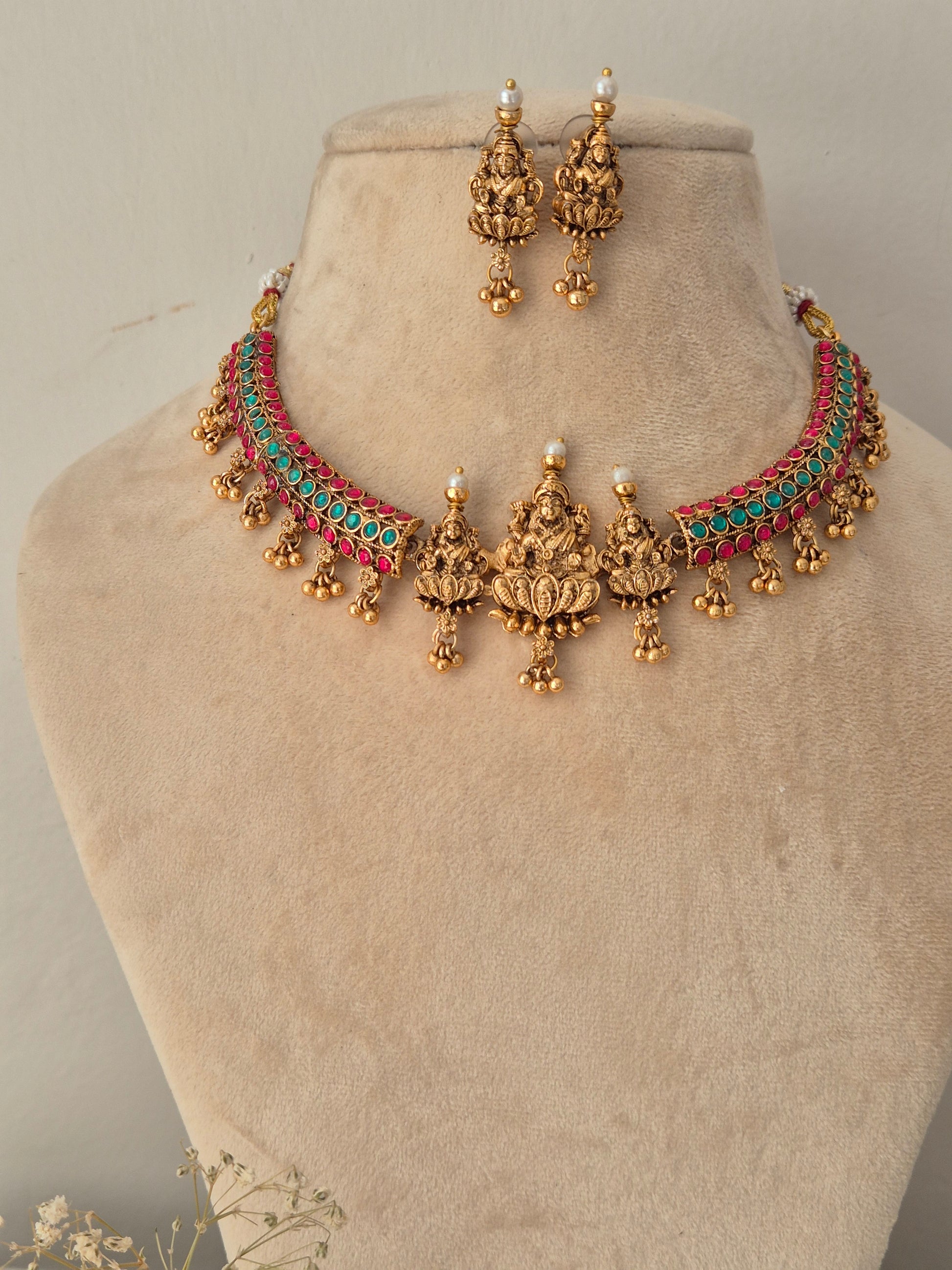 Gold-toned traditional Indian jewelry set including a choker necklace and matching earrings, decorated with red and green stones and gold bell details, by naitri.in